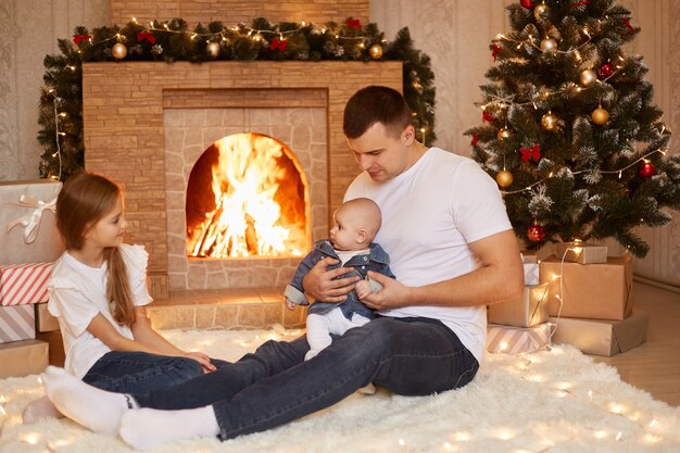 Innenaufnahme eines jungen erwachsenen kaukasischen Vaters, der mit zwei Töchtern in der Nähe von Kamin und Weihnachtsbaum auf dem Boden sitzt und gemeinsam Winterferien feiert.