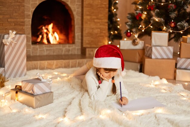 Innenaufnahme eines kleinen süßen Mädchens mit weißem Pullover und rotem Hut, das auf einem weichen Teppich in einem festlich dekorierten Raum auf dem Boden liegt und einen Brief an den Weihnachtsmann schreibt.