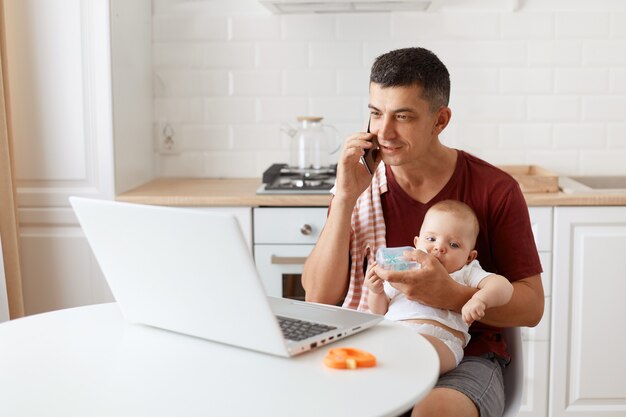Innenaufnahme eines Mannes, der ein burgunderfarbenes Casual-T-Shirt mit einem Handtuch auf der Schulter trägt, sich um das Baby kümmert und von zu Hause aus online arbeitet, telefoniert und seinem Kind Wasser gibt.