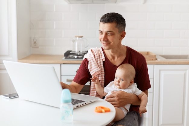 Innenaufnahme eines Mannes, der ein burgunderfarbenes Casual-T-Shirt mit einem Handtuch auf der Schulter trägt, sich um das Baby kümmert und von zu Hause aus online arbeitet und lächelnd auf den Notebook-Bildschirm schaut.