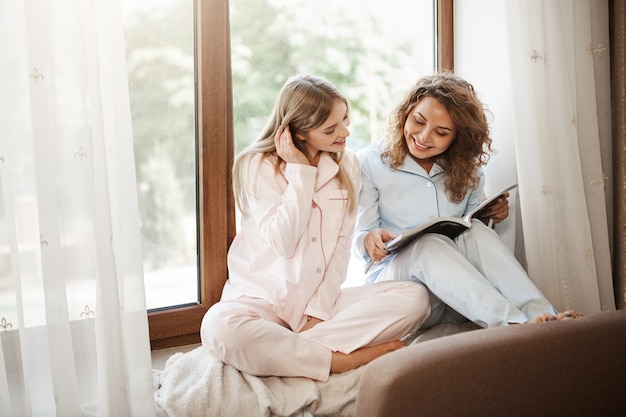 Innenaufnahme von entspannten glücklichen kaukasischen Schwestern, die zu Hause auf dem Fensterbrett in niedlicher Nachtwäsche sitzen, Artikel in der Zeitschrift lesen, den neuesten Trend in der Modebranche diskutieren oder neue Kleidung auswählen