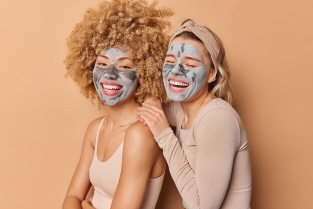 Innenaufnahme von glücklichen jungen hübschen Frauen wenden Tonmasken an Lächeln breit gekleidet lässig stehen eng beieinander vor braunem Hintergrund Pose Gesichtsbehandlungen Schönheits- und Hautpflegekonzept