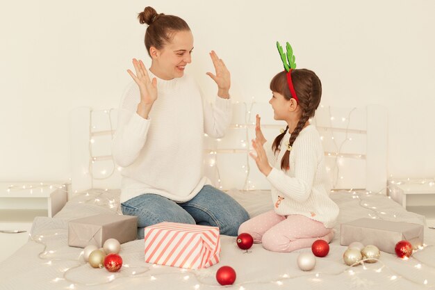 Innenaufnahme von optimistischer Mutter und Tochter, die weiße Pullover im lässigen Stil tragen, die auf dem Bett sitzen, einander fünf geben, Weihnachten feiern, ein frohes neues Jahr.