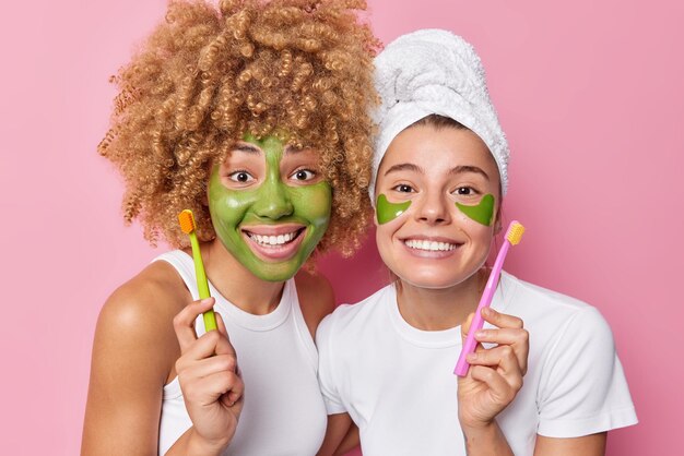 Innenaufnahmen positiver junger Frauen wenden grüne Schönheitsmaske an und Hydrogel-Patches halten Zahnbüsche und kümmern sich um Zähne, die in Freizeitkleidung gekleidet sind, die über rosafarbenem Hintergrund isoliert ist Mundhygienekonzept