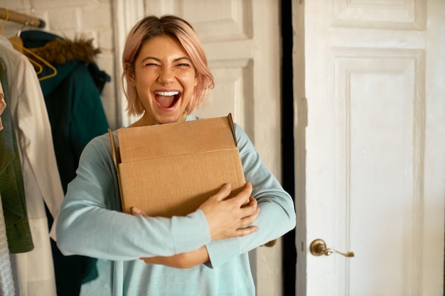 Innenbild der glücklichen fröhlichen jungen Frau, die Pappkarton hält, der zu ihrer Wohnung geliefert wird, Aufregung ausdrückt, Paket auspacken geht