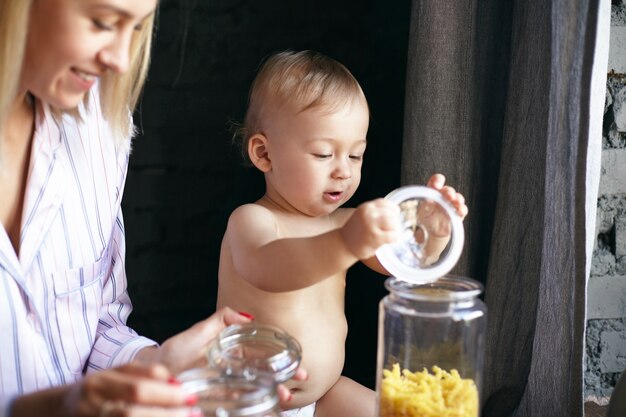 Innenbild des entzückenden glücklichen Kleinkindes in der Windel, die mit dem Deckel der Glasflasche in der Küche spielt, seine schöne junge Mutter, die neben ihm sitzt und breit lächelt. Selektiver Fokus auf Babys Gesicht