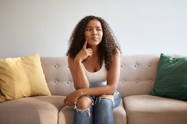 Innenbild des modischen niedlichen Mädchens mit afrikanischem Aussehen, das isoliert auf grauem Sofa mit gelben und grünen Kissen sitzt, die zweifelhaften nachdenklichen Blick haben, Kinn berühren und Hmm sagen, lassen Sie mich denken