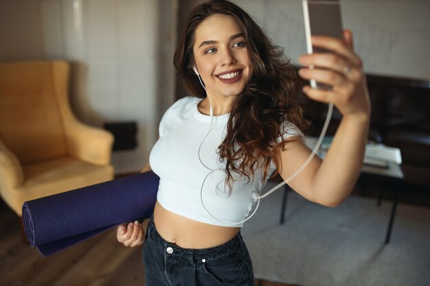 Innenbild des niedlichen charmanten Teenager-Mädchens im Erntedach und in den Jeans, die Yogamatte und Smartphone halten, posiert für Selfie in Kopfhörern, glücklich lächelnd. Aktiver Lebensstil, Training und moderne Geräte