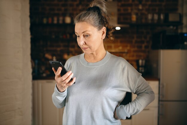 Innenbild einer ernsthaften grauhaarigen Rentnerin in Freizeitkleidung, die Tage zu Hause wegen sozialer Distanzierung verbringt und Weltnachrichten liest, während sie mit WLAN im Internet auf dem Handy surft