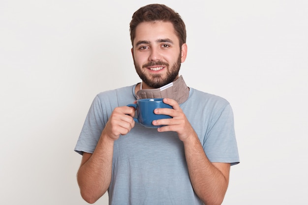 Innenfoto des fröhlichen aufrichtigen jungen Mannes, der Bart hat und direkt Tasse mit Kaffee hält und angenehmes Lächeln hat