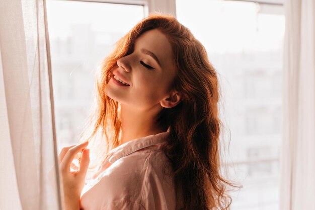 Innenfoto eines sinnlichen rothaarigen Mädchens, das mit geschlossenen Augen lächelt Hübsche, fröhliche Frau, die in der Nähe des Fensters posiert