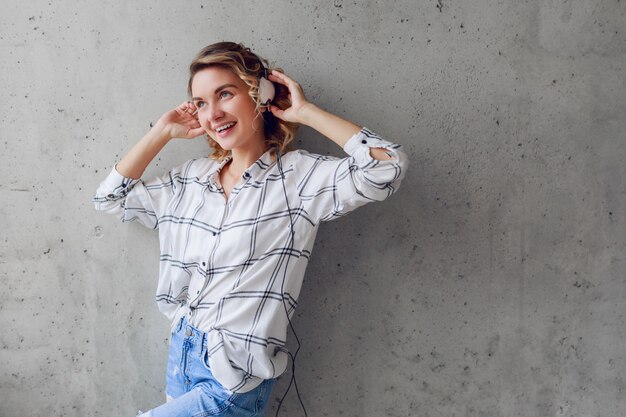 Innenlebensstilporträt der glücklichen begeisterten Frau, die Musik auf Stuhl auf grauem Stadtwandhintergrund hört.