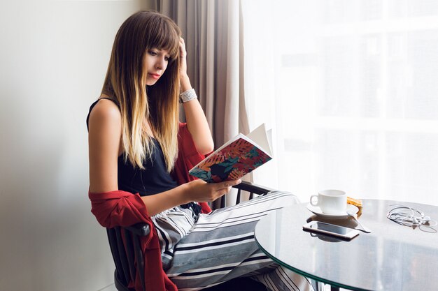 Innenlebensstilporträt der hübschen brünetten Frau, die Buch liest, auf Stuhl sitzt und Kaffee im sonnigen Frühlingsmorgen trinkt. Frühstückszeit.