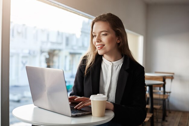 Innenporträt der attraktiven europäischen Frau, die im Café sitzt, Kaffee trinkt und in Laptop tippt, glücklich und erfreut ist.