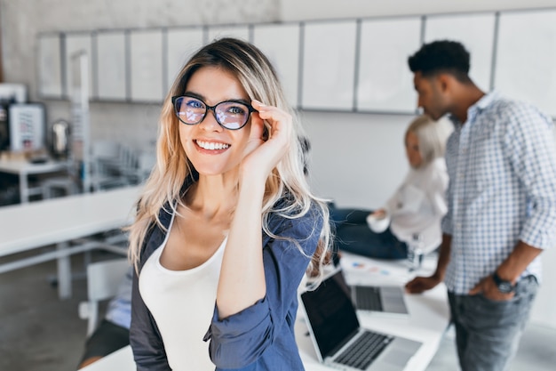 Innenporträt der aufgeregten Studentin in Brille und grauer Jacke. Attraktive Mitarbeiterin, die im Büro posiert und mit Kollegen lacht.