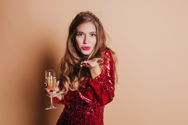 Innenporträt der entzückenden europäischen Frau im roten Kleid, das Luftkuss sendet und Glas Champagner hält