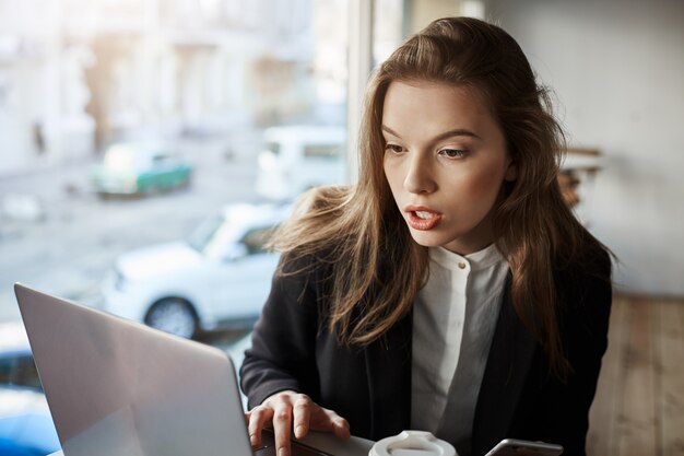 Innenporträt der gestörten und verwirrten stilvollen Frau, die im Café sitzt, mit Laptop arbeitet und Bildschirm mit überraschtem Ausdruck betrachtet