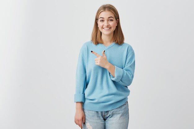 Innenporträt der schönen hübschen jungen Frau mit hellem Haar, das lässigen blauen Pullover und Jeans mit angenehmem Lächeln trägt, das mit Finger auf Kopierraum für Ihre Anzeige oder Text zeigt.