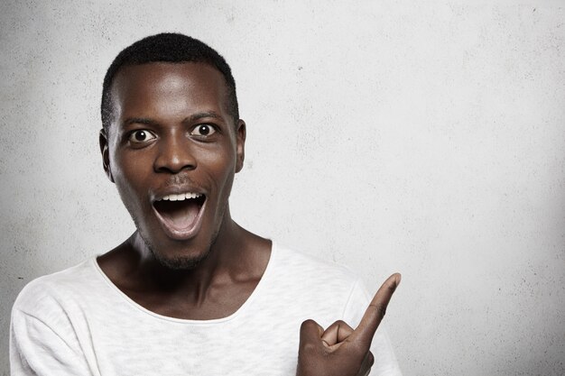 Innenporträt des afrikanischen Mannes, der weißes T-Shirt mit erstauntem und überraschtem Blick trägt, Mund weit öffnet und mit seinem Zeigefinger auf leere Wand anzeigt.