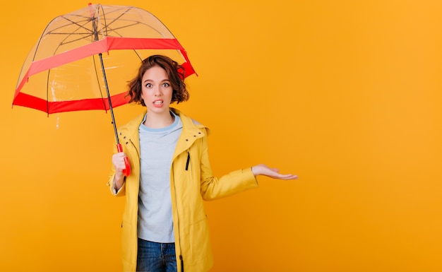 Kostenloses Foto innenporträt des modischen mädchens im regenmantel, der regenschirm auf gelber wand hält. lockige junge dame mit sonnenschirm, der erstaunen ausdrückt.