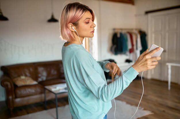 Innenporträt des stilvollen Teenager-Mädchens mit rosa Bob-Frisur, die schöne Zeit zu Hause genießt und Smartphone hält