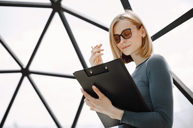 Innenporträt einer jungen Geschäftsfrau, die in einem Café steht und auf einem Papierhalter schreibt. Blondes Mädchen mit Sonnenbrille