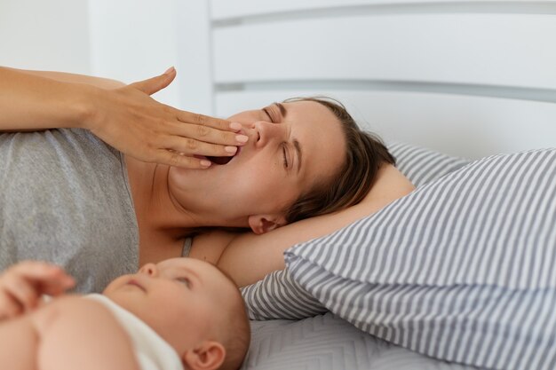 Innenporträt einer müden Mutter, die mit einem kleinen Mädchen oder Jungen im Bett liegt, gähnt, den Mund mit der Handfläche bedeckt, schläfrig aussieht, Energiemangel und schlaflose Nächte hat.