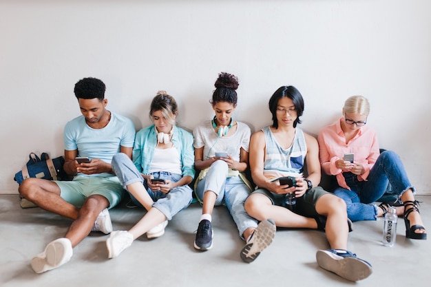 Innenporträt von internationalen Studenten, die auf die Prüfung warten und ihre Telefone benutzen. Jungen und Mädchen sitzen mit gekreuzten Beinen auf dem Boden und halten Geräte in Händen.