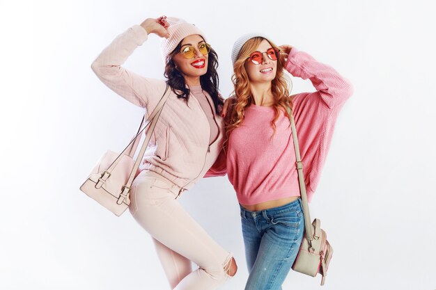 Innenstudio Bild von zwei Mädchen, glücklichen Freunden in stilvollen rosa Kleidung und Hut Rechtschreibung lustig zusammen. Weißer Hintergrund. Trendy Hut und Brille. Frieden zeigen.