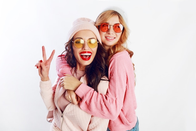 Innenstudio Bild von zwei Mädchen, glücklichen Freunden in stilvollen rosa Kleidung und Hut Rechtschreibung lustig zusammen. Weißer Hintergrund. Trendy Hut und Brille. Frieden zeigen.