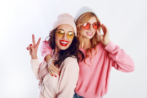 Innenstudio Bild von zwei Mädchen, glücklichen Freunden in stilvollen rosa Kleidung und Hut Rechtschreibung lustig zusammen. Weißer Hintergrund. Trendy Hut und Brille. Frieden zeigen.