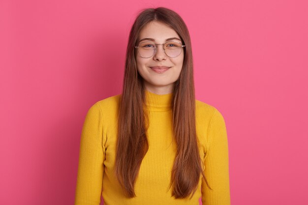 Innenstudioaufnahme der attraktiven schönen hübschen jungen Frau, die Brillen und gelbes Sweatshirt trägt, langes blondes Haar hat, das lokal über rosa Wand aufwirft. Menschen- und Emotionskonzept.