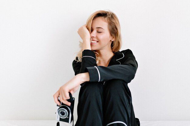 Innenstudioaufnahme der reizenden glücklichen Frau schloss Augen und smiing, während auf dem Bett über weißer Wand mit Retro-Kamera sitzend