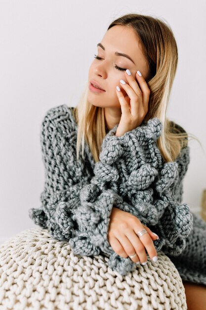 Innenstudioaufnahme der reizenden sinnlichen Frau mit den blonden Haaren und den geschlossenen Augen, die ihr Gesicht berührend Strickpullover berühren, der im weißen Raum posiert