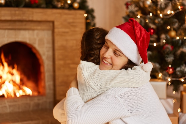 Innenstudioaufnahme von Mutter und ihrer kleinen Tochter, die sich im festlichen Raum in der Nähe von Kamin und Weihnachtsbaum umarmen und sich zu Silvester gratulieren.