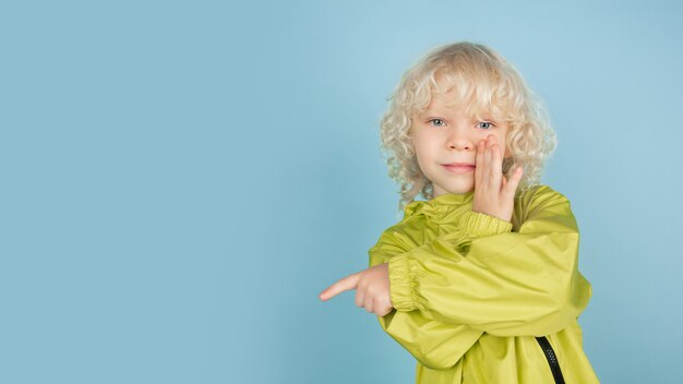 Insgeheim zeigen. Porträt des schönen kaukasischen kleinen Jungen lokalisiert auf blauer Studiowand. Blondes lockiges männliches Model