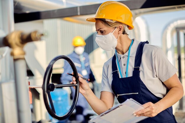 Inspektorin mit Gesichtsmaske untersucht Maschinenventil in einer Industrieanlage im Freien