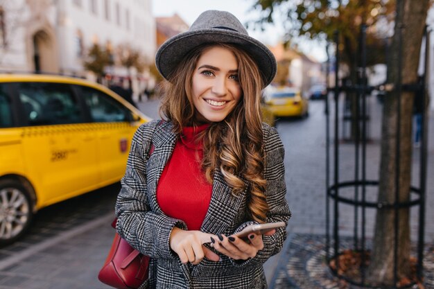 Inspirierte blauäugige Geschäftsfrau, die mit Telefon auf der Straße posiert