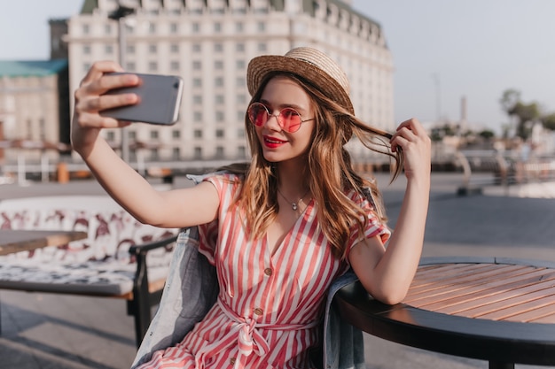 Inspirierte europäische Dame im Strohhut, die mit ihren Haaren spielt und Selfie macht. Foto im Freien des liebenswerten weißen Mädchens im gestreiften Kleid, das Foto von sich auf Stadt nimmt.