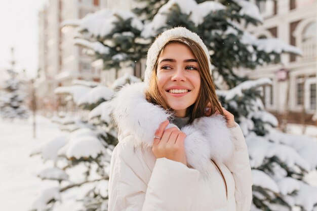 Inspirierte europäische Dame trägt weiße Winterkleidung, die Naturansichten genießt. Außenporträt des atemberaubenden kaukasischen weiblichen Modells lächelnd