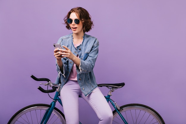 Inspirierte Frau in der Freizeitkleidung, die Telefonbildschirm betrachtet. Freudiges weißes Mädchen in der dunklen Sonnenbrille, die Smartphone mit Fahrrad verwendet.