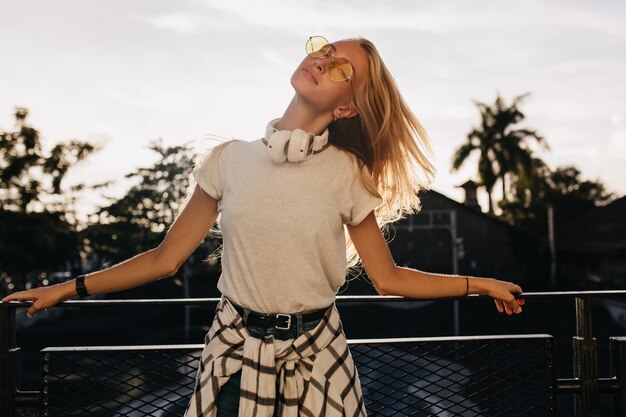 Inspirierte Frau in der gelben Sonnenbrille, die mit den Augen geschlossen auf Himmelhintergrund aufwirft.