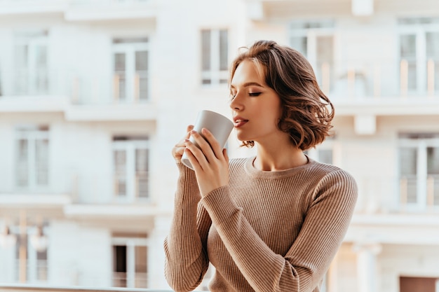 Inspirierte weiße Dame mit lockiger Frisur, die Tee trinkt. Wunderschöne junge Frau, die Kaffee im kalten Herbstmorgen genießt.