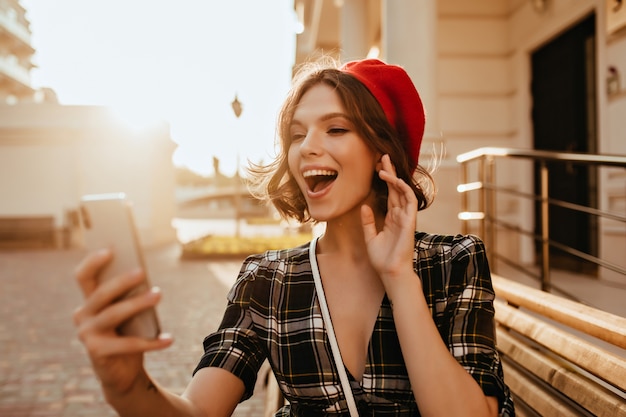 Inspirierte weiße Frau mit kurzem Haarschnitt, die Spaß im Herbsttag hat. Foto im Freien des faszinierenden französischen weiblichen Modells in der Baskenmütze, die Smartphone hält.