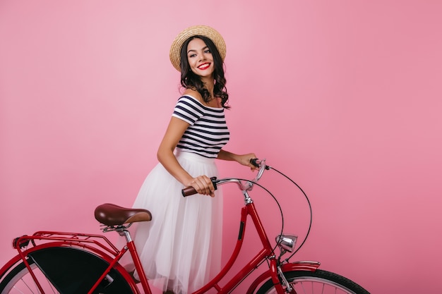 Inspirierte wohlgeformte Frau, die mit Fahrrad steht und wegschaut. Glückseliges braunhaariges Mädchen im Hut, das Innen-Fotoshooting genießt.