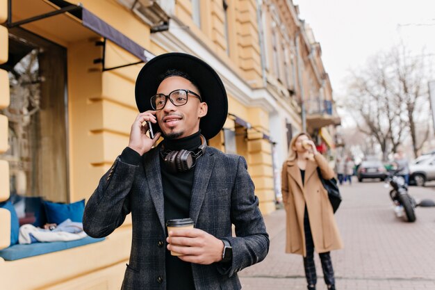 Inspirierter afrikanischer Junge, der Kaffee auf der Straße trinkt. Außenporträt des sorglosen schwarzen männlichen Modells, das Latte genießt und am Telefon spricht.