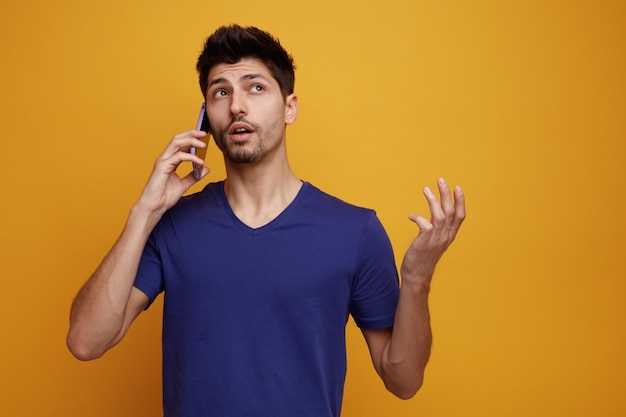 Inspirierter junger gutaussehender Mann, der am Telefon spricht und nach oben schaut und leere Hand auf gelbem Hintergrund zeigt
