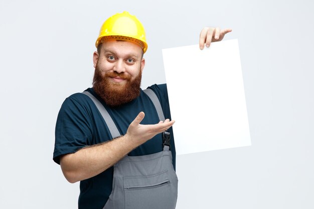Inspirierter junger männlicher Bauarbeiter, der Schutzhelm und Uniform trägt und in die Kamera schaut, die Papier zeigt, um Kamera zu zeigen, die mit der Hand auf Papier zeigt, isoliert auf weißem Hintergrund