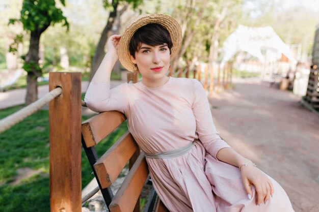 Inspiriertes braunhaariges Mädchen, das auf Holzbank ruht und auf Freunde wartet, um Zeit zusammen im Freien zusammen zu verbringen