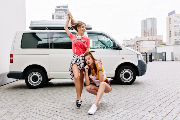 Inspiriertes brünettes Mädchen im gelben Hemd sitzt mit gekreuzten Beinen neben weißem Auto, während ihre Freundin mit langen Haaren spielt